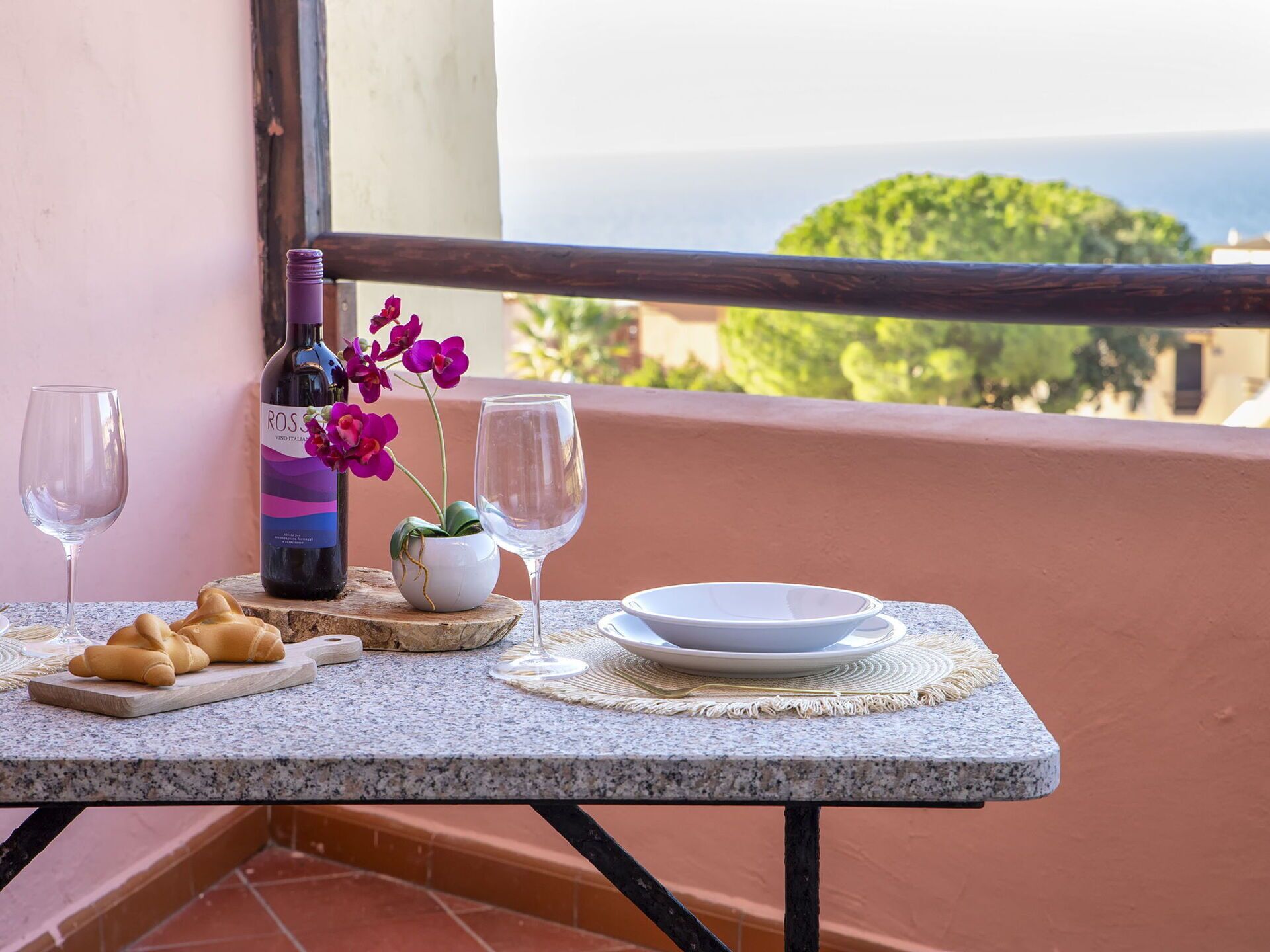 Un balcon avec une table dressée pour deux personnes, comprenant une bouteille de vin, deux verres, une assiette, un bol, une petite plante en pot et des croissants.