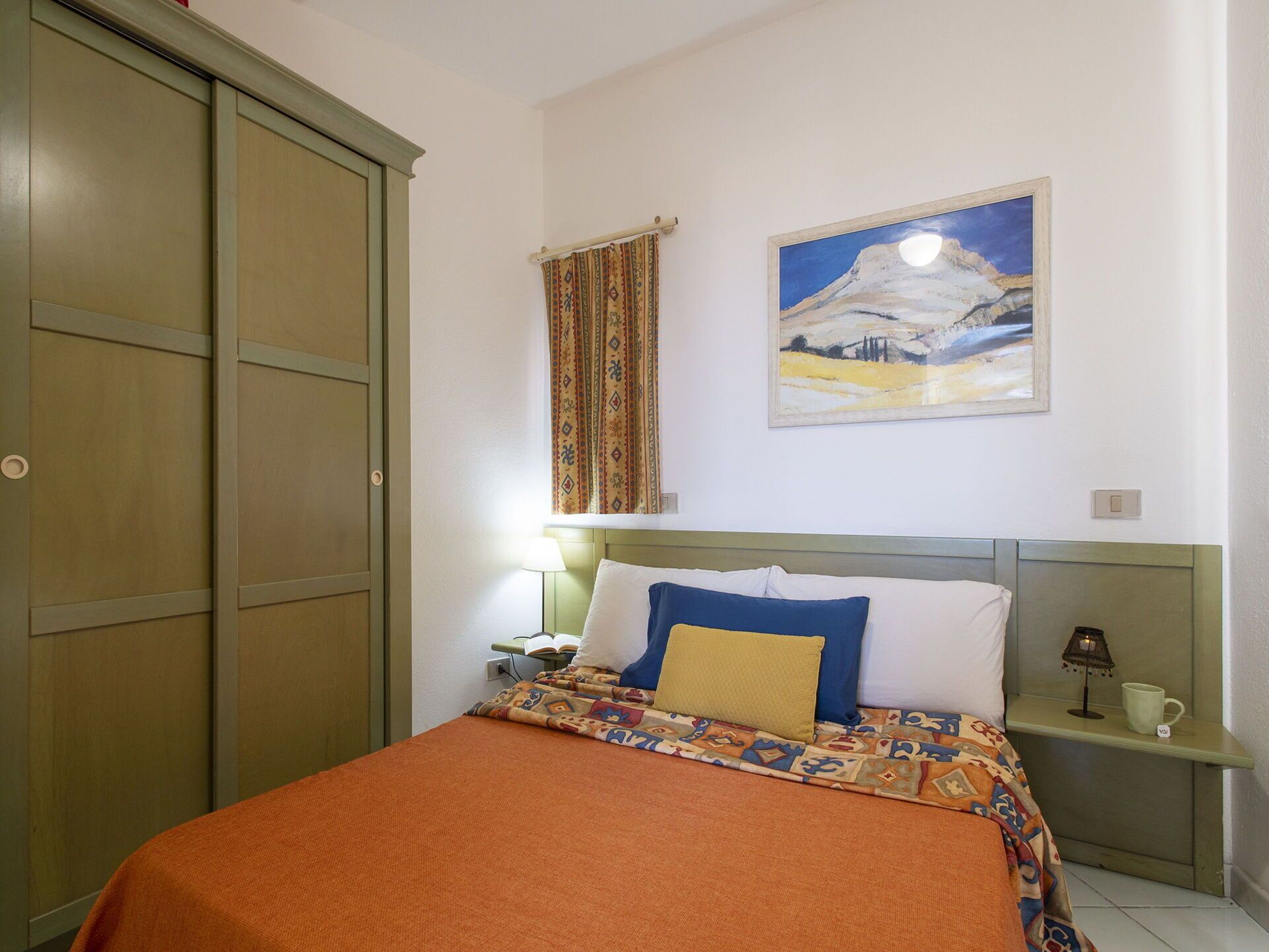 A bedroom with a bed, a nightstand, a lamp, a painting, and a wardrobe.