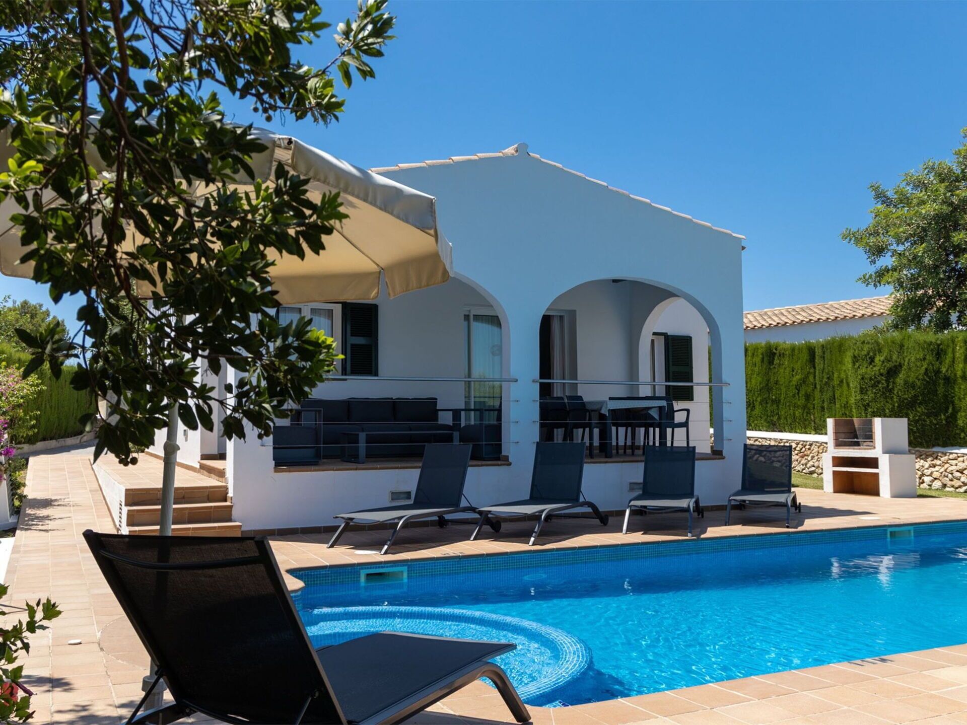 A modern outdoor pool area with a white building, lounge chairs, and a barbecue area.