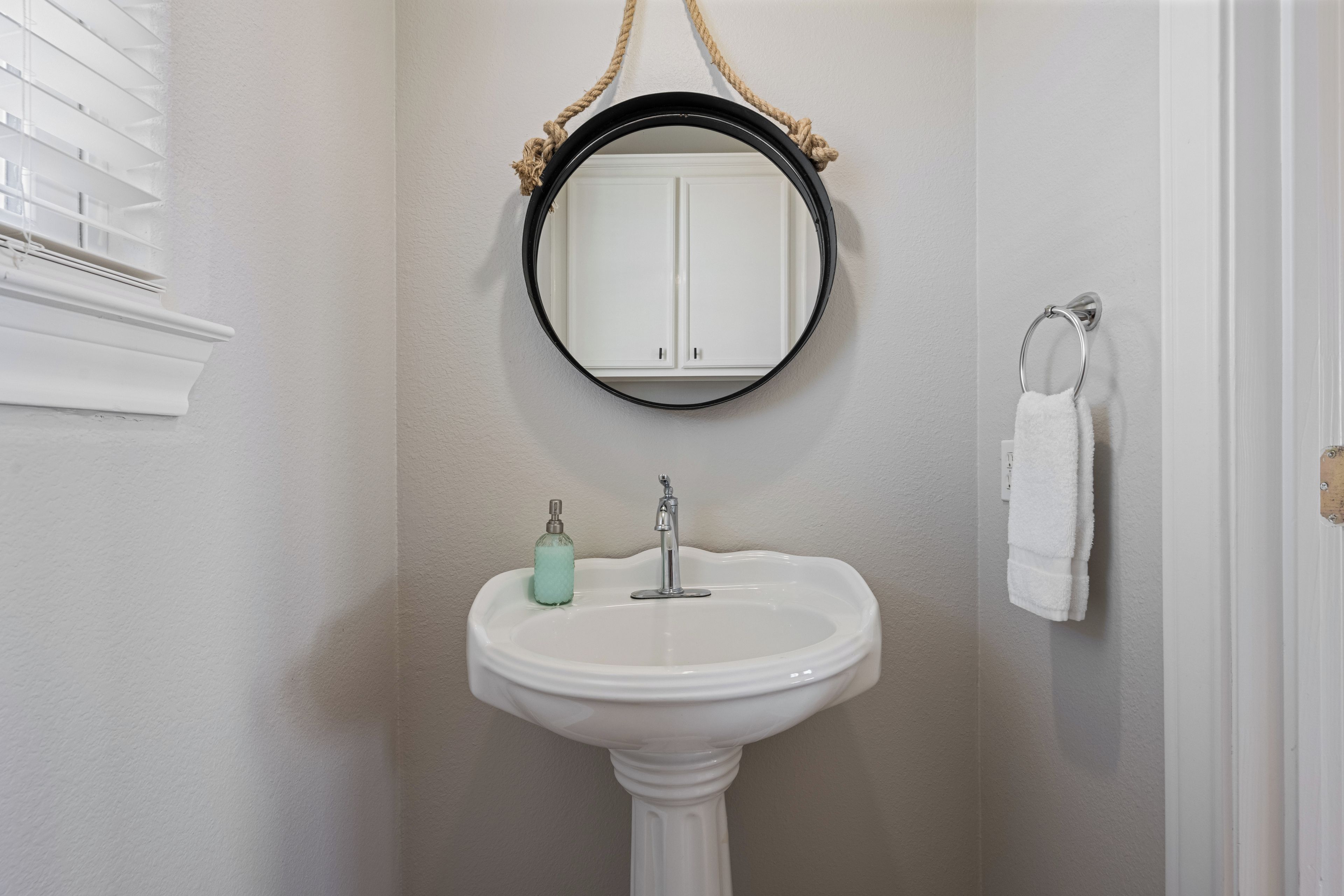 A bathroom with a white pedestal sink, a round mirror with a rope handle, and a towel rack with a white towel.