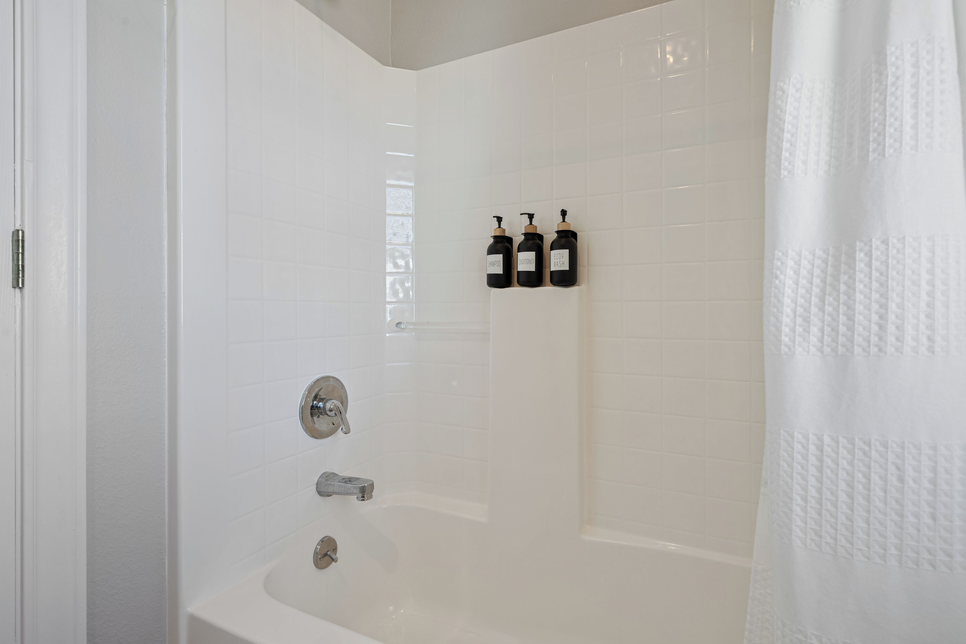 Ein helles Badezimmer mit einer Badewanne, einer Dusche und drei schwarzen Flaschen mit den Aufschriften 'shampoo', 'conditioner' und 'body wash' an der Wand.