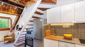 Eine moderne Küche mit Edelstahl-Kühlschrank, Backofen und Geschirrspüler. Eine Holztreppe führt auf eine obere Ebene, und ein Fenster bietet Blick ins Grüne.