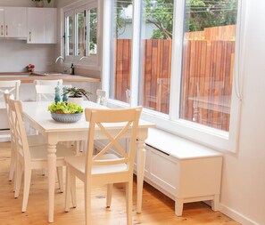 A modern kitchen with white cabinetry, a dining table with four chairs, a bench, and a window overlooking a wooden fence. - Peaceful Places - Surf Side, Linen, Pets, Wifi (Inverloch)