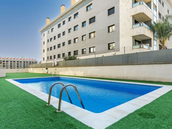 Una piscina con una escalera de metal, rodeada de césped artificial, y un edificio de varios pisos con balcones al fondo.