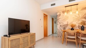 A modern living room with a flat-screen TV on a wooden cabinet, a dining area with a table and chairs, and a wall with a peacock feather pattern. - Beautiful apartment for 4 people with pool, WIFI, A/C, TV and terrace (Lloret De Mar)