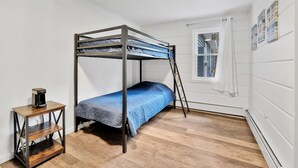 Una cama litera con colchón azul, una mesita de madera con cafetera y una ventana con cortinas.