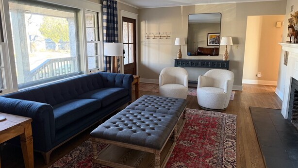 Een zithoek met een blauwe bank, twee fauteuils, een grijze poef, een houten salontafel, een vloerkleed met patroon, een open haard en een grote spiegel aan de muur.