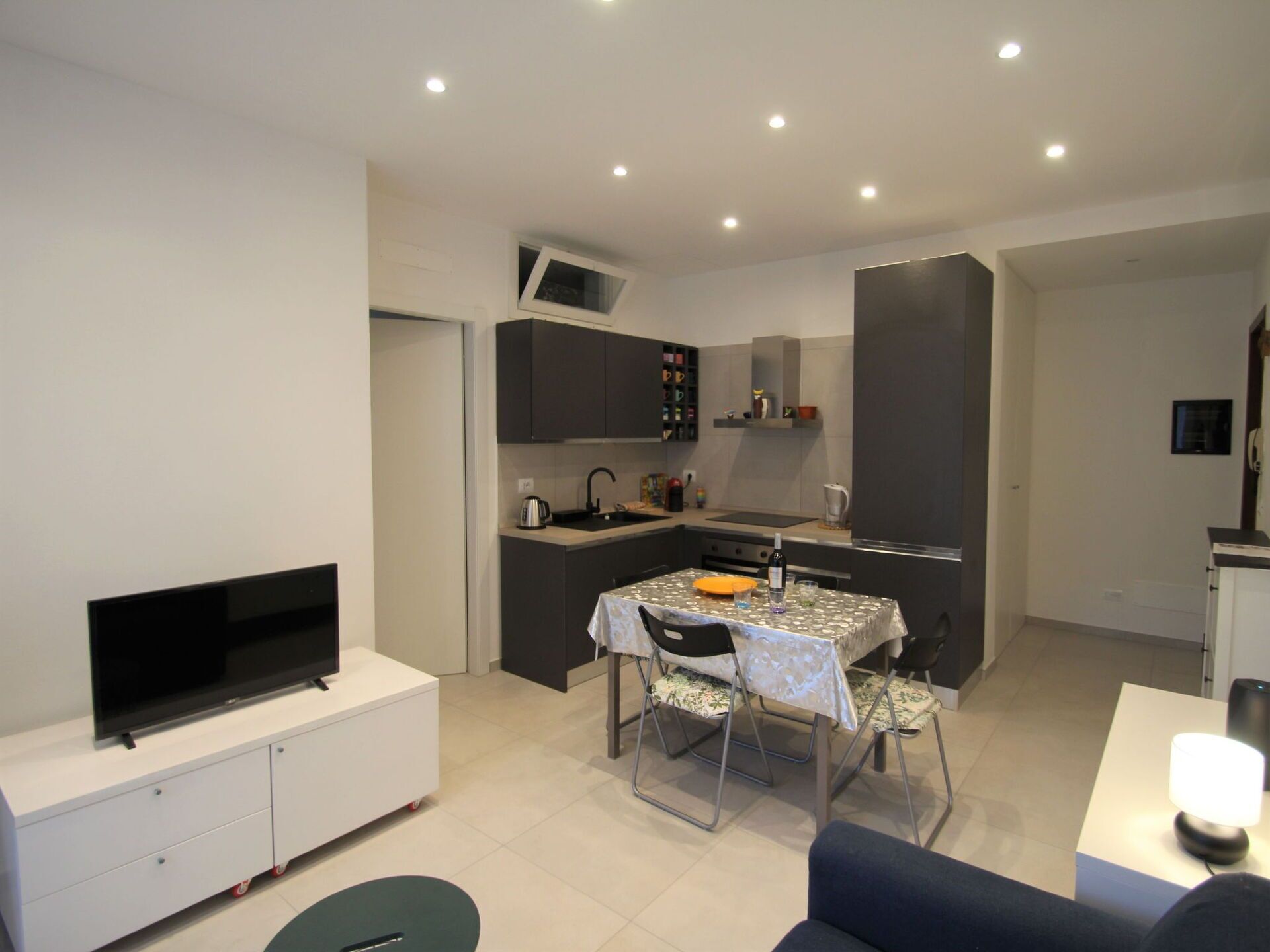 A compact living space with a kitchenette, a dining area with a table and chairs, a sofa, a TV on a white cabinet, and a small table with a lamp.