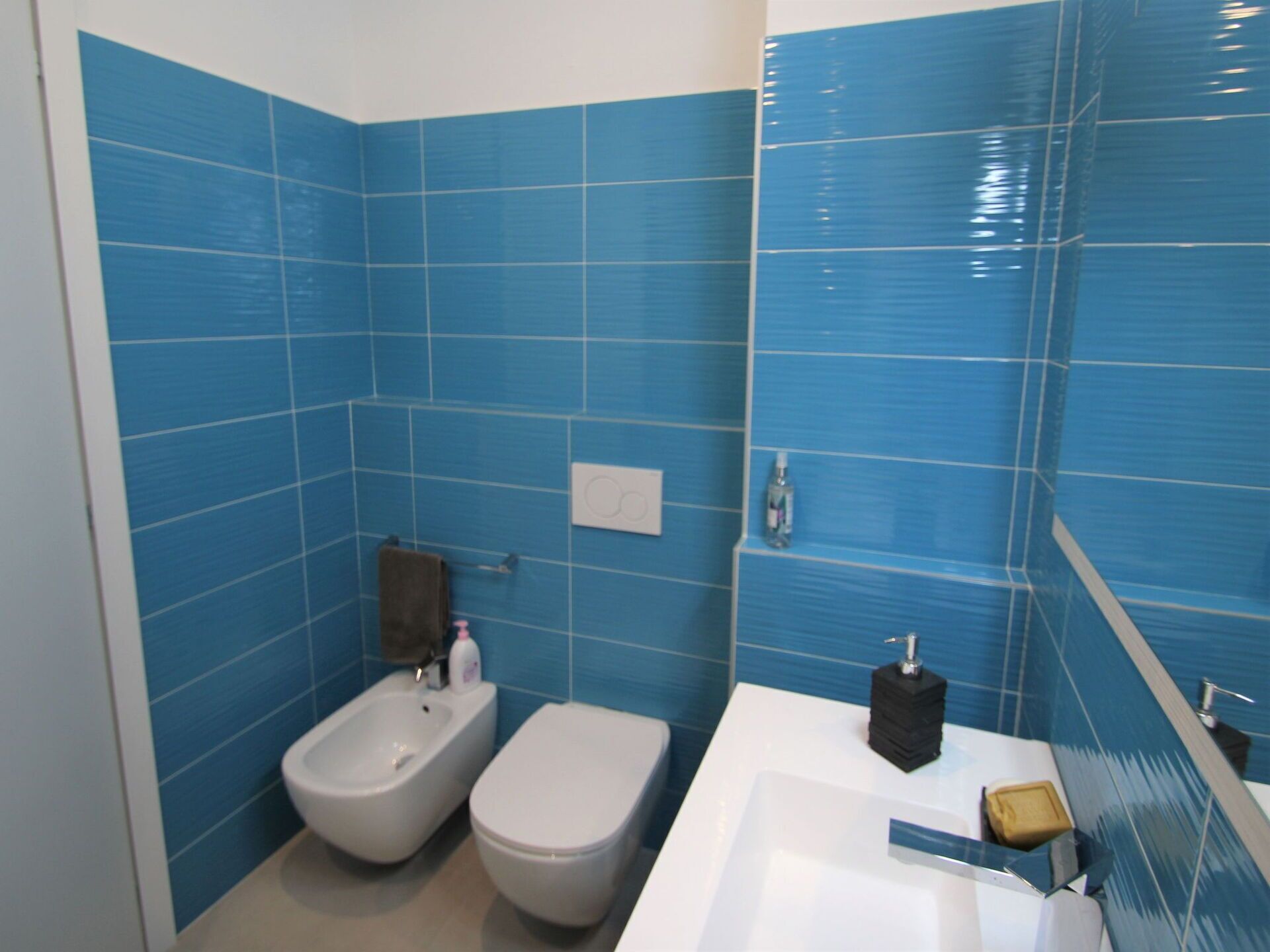 A bathroom with blue tiles, a bidet, a toilet, a sink, and a soap dispenser.