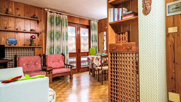 A cozy living room with a wooden floor, a sofa, a chair, and a dining table. There is a bookshelf with books and decorative items.