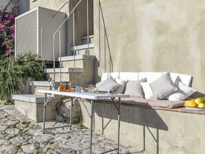 A stone patio with a white table and a cushioned bench against a concrete wall. - Beautiful apartment for 2 guests with A/C, WIFI, TV, terrace and pets allowed (Marciana)