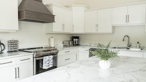 A modern kitchen with white cabinets, a stainless steel stove, a coffee maker, and a marble countertop.