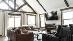 Un salón amplio con techo alto, ventanas grandes, un televisor de pantalla plana, sofás de cuero y un piano.