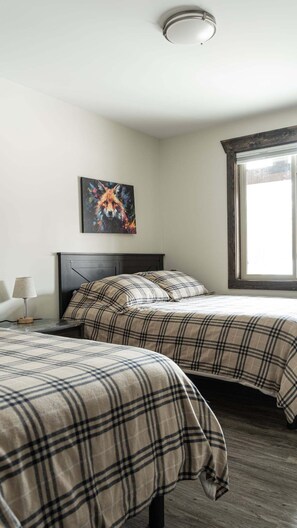 A bedroom with two beds, a painting on the wall, a lamp, and a window with curtains. - Highmark Cabin Rental (Valemount)