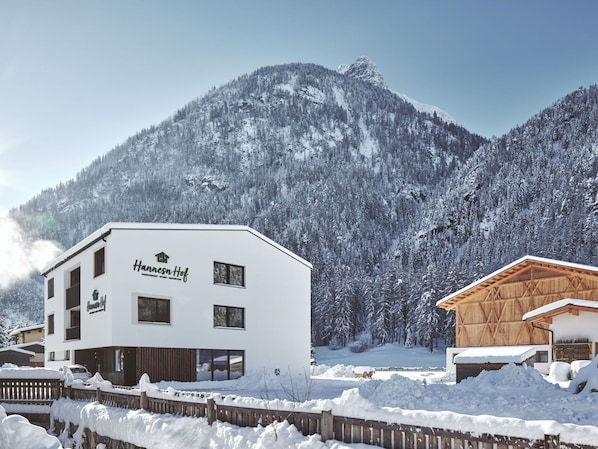 A white building with "Hansenhof" signage, surrounded by snow-covered mountains and a wooden fence.