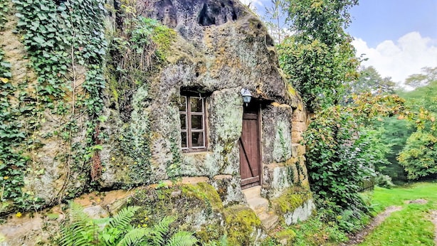 Una casa de campo rústica de piedra, con una pequeña ventana y puerta, rodeada de exuberante follaje y helechos.