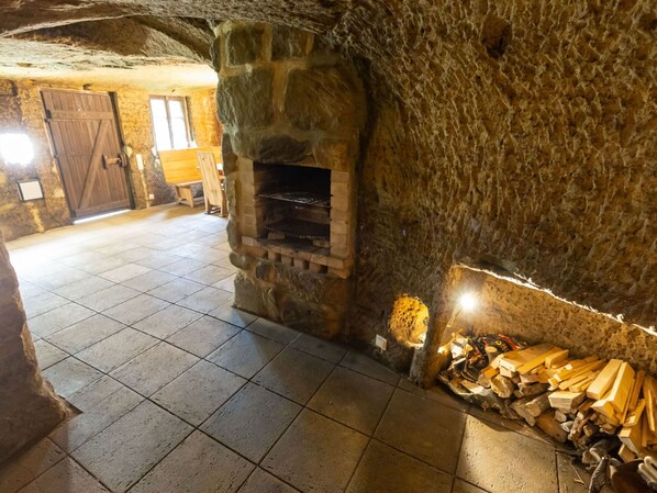 Una habitación de paredes de piedra con chimenea, suelo de baldosas y una pila de leña.