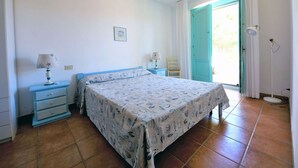 Un dormitorio con cama, dos mesitas de noche con lámparas, una silla, un cuadro en la pared y una puerta que da a un balcón.