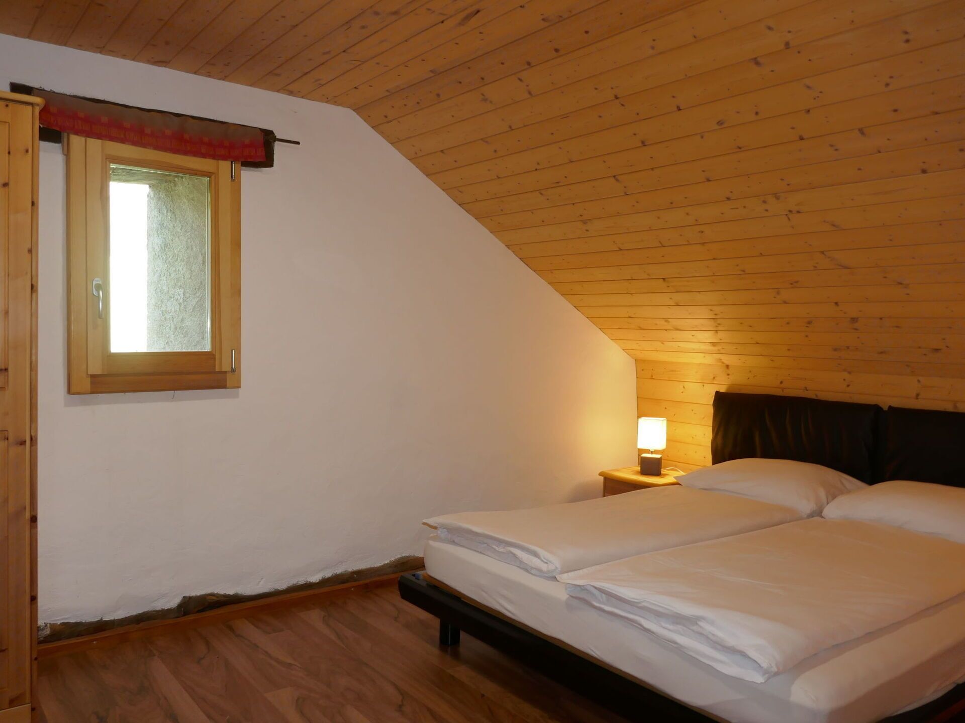 A bedroom with a wooden ceiling, a window with a red curtain, a bedside table with a lamp, and a bed with white bedding.