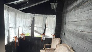 En bänk i gammaldags stil står i ett svagt upplyst rum med träväggar och ett fönster med spetsgardiner som är knutna med orangefärgade band.