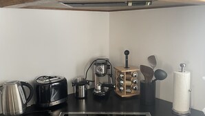 Ett kök med en svart induktionshäll, en kaffebryggare, en brödrost, en vattenkokare och en hylla i trä ovanför.