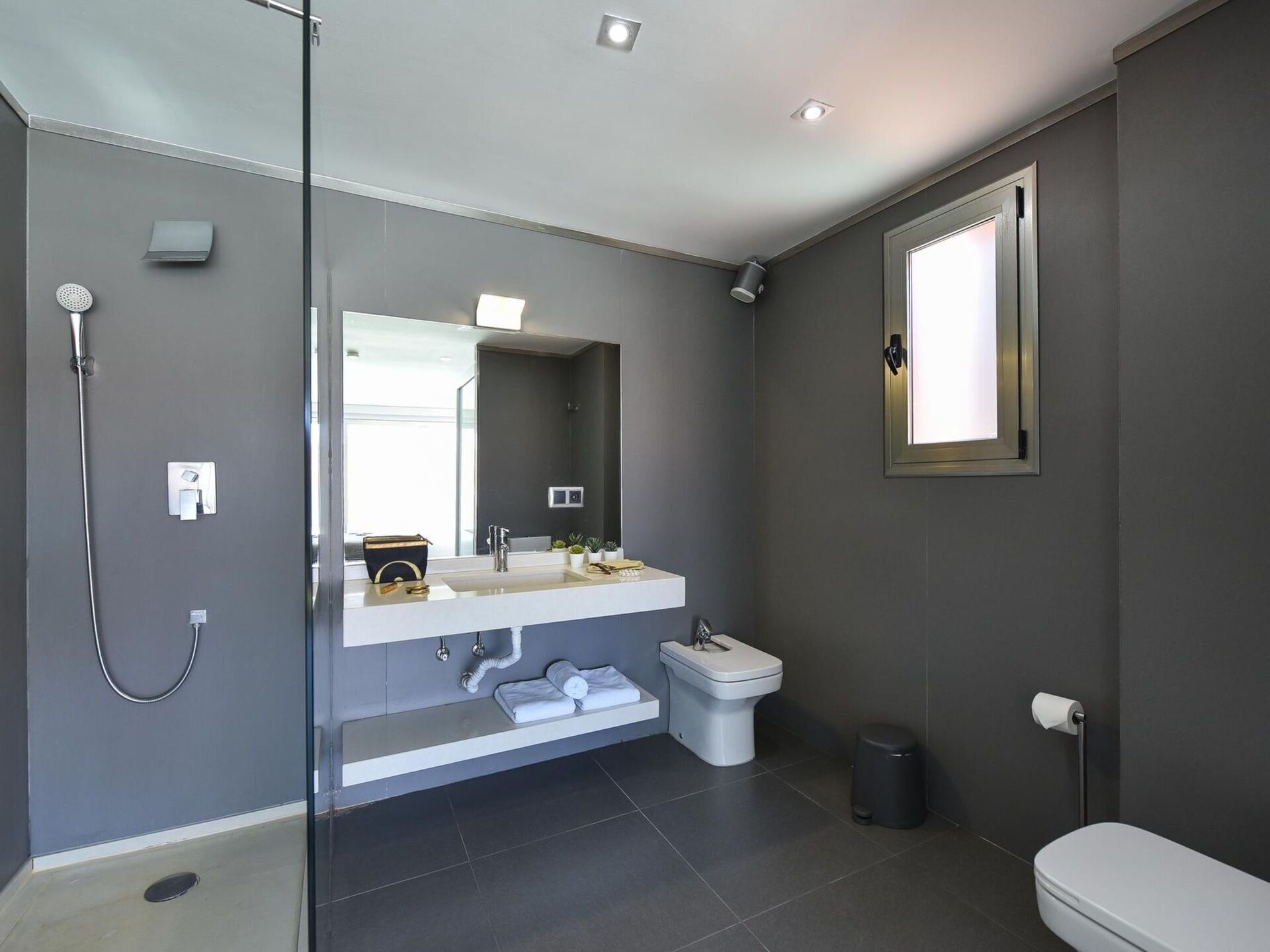 Ein modernes Badezimmer mit Duschkabine aus Glas, einem weißen Waschbecken mit Spiegel darüber, einer Toilette und einem Mülleimer.