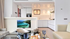A modern living room with a flat-screen TV, a dining table with a breakfast setup, a sofa, and a kitchen area with white cabinets and a framed picture on the wall.