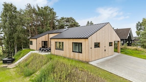 A modern wooden house with a sloped roof, a patio with outdoor seating, and a driveway leading to the entrance.
