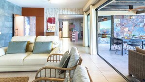 Un vaste espace de vie comprenant un canapé, un fauteuil en osier et une table à manger avec des chaises. Une porte-fenêtre coulissante donne accès à un patio extérieur.