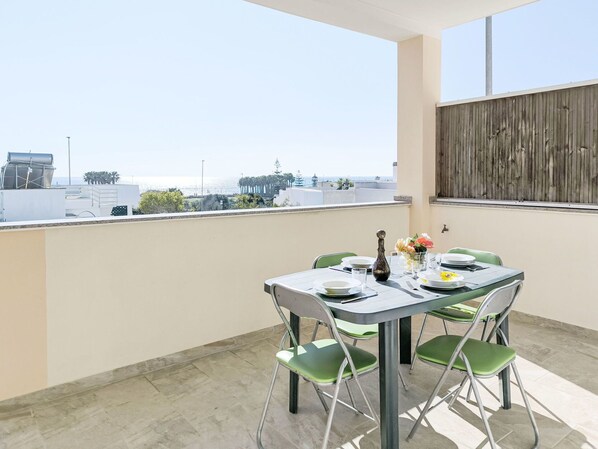 A rooftop terrace with a table set for two, green chairs, and a view of the cityscape and sea.