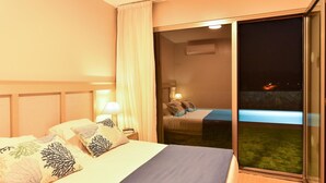 A hotel room with a bed, a nightstand, a lamp, and a sliding glass door leading to a balcony with a view of the night sky and distant lights.