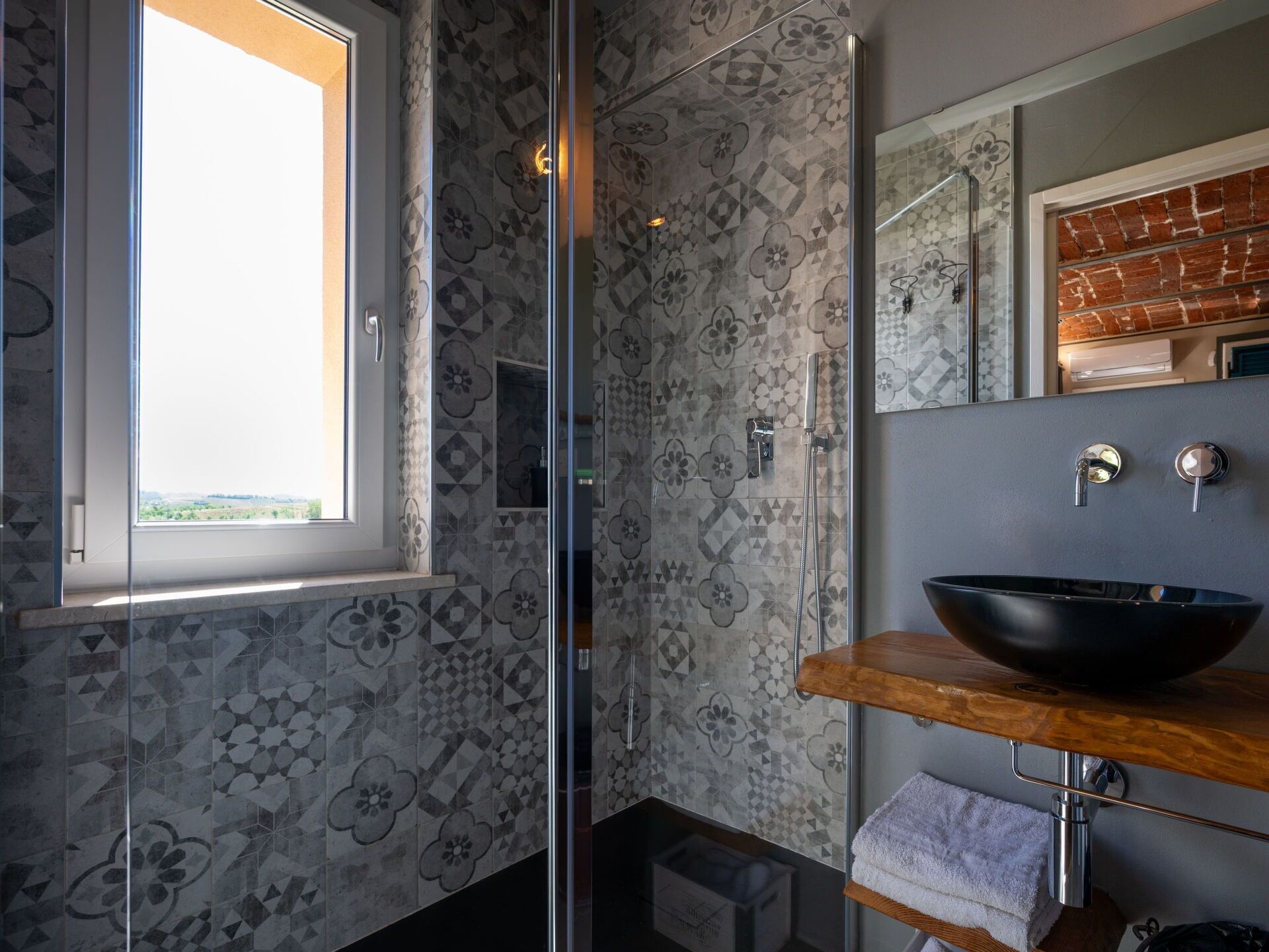 Un baño con un salpicadero de azulejos geométricos, un ventanal grande, una cabina de ducha de cristal, un tocador de madera con un lavabo negro y un espejo sobre el lavamanos.