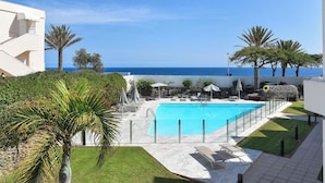 A pool area with lounge chairs, a clear view of the sea, and a white building. - Beautiful apartment for 2 people with A/C, pool, WIFI, TV and balcony (Playa Del Ingles)