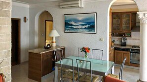 A dining area with a table and chairs, a lamp, a painting of the ocean, and a kitchen in the background. - Nice apartment in Privlaka (Privlaka)