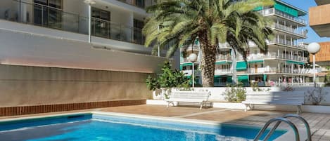 A swimming pool with a palm tree and benches, surrounded by multi-story buildings with balconies.