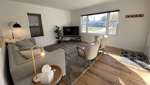 Una sala de estar con un sofá, sillas, un televisor, una lámpara y una ventana con vistas a casas.