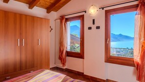 A bedroom with a wooden wardrobe, a bed with a striped blanket, a window with a view of mountains and a lake, and a ceiling with exposed beams. - Wonderful private villa for 5 people with WIFI, TV and terrace (Peglio)