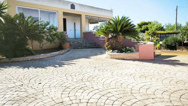 Un edificio con un letrero que dice "Villa Cosimo", que presenta un patio empedrado con un sendero de piedra, palmeras y una pérgola con asientos.