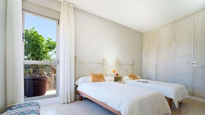 A bedroom with two beds, a window with curtains, a small table with a lamp, and a view of greenery outside.