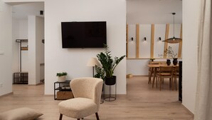 A modern living room with a flat-screen TV mounted on the wall, a comfortable armchair, a small side table with a lamp and a potted plant, and a dining area in the background. - Amazing home in Rizvanusa (Gospic)