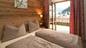 A wooden room with two beds, a painting on the wall, and a view of a snowy landscape through a glass door.