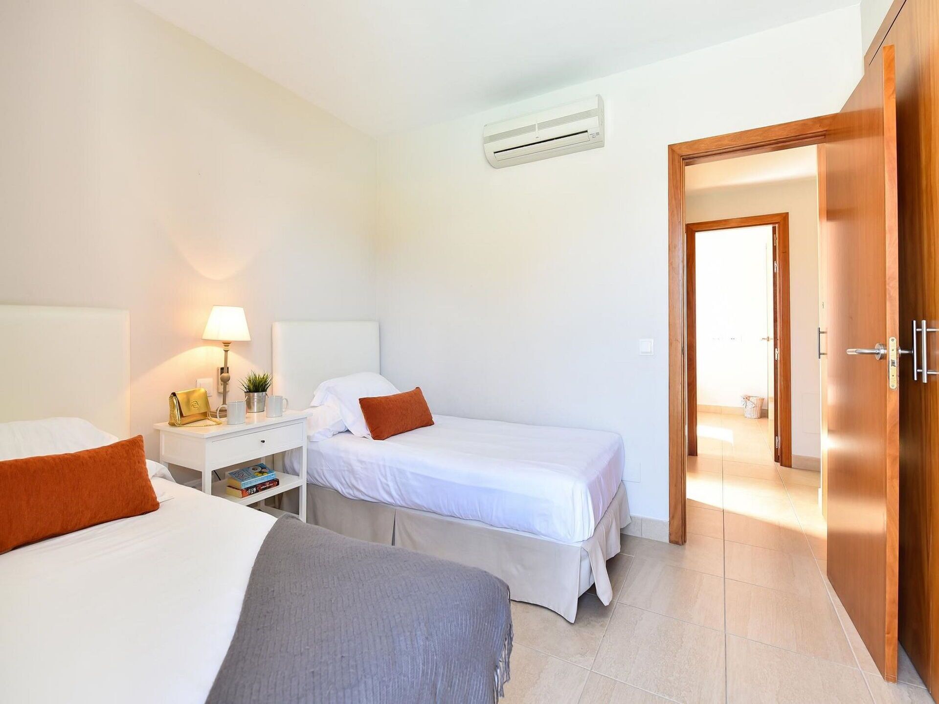 Camera d'albergo con due letti, un comodino con lampada, una piccola pianta e una porta che dà su un'altra stanza.