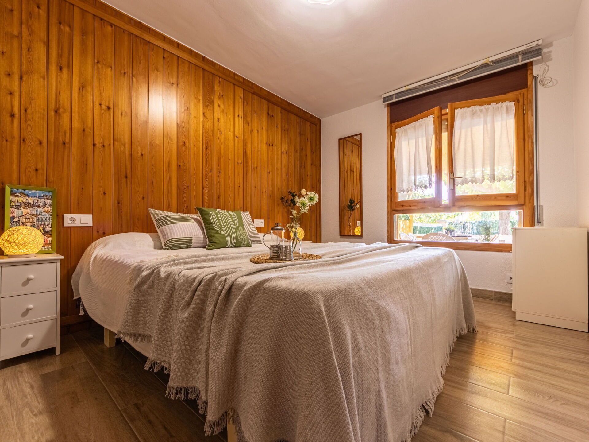 A bedroom with a wooden paneled wall, a bed with a white bedspread, a bedside table with a lamp, and a window with curtains.