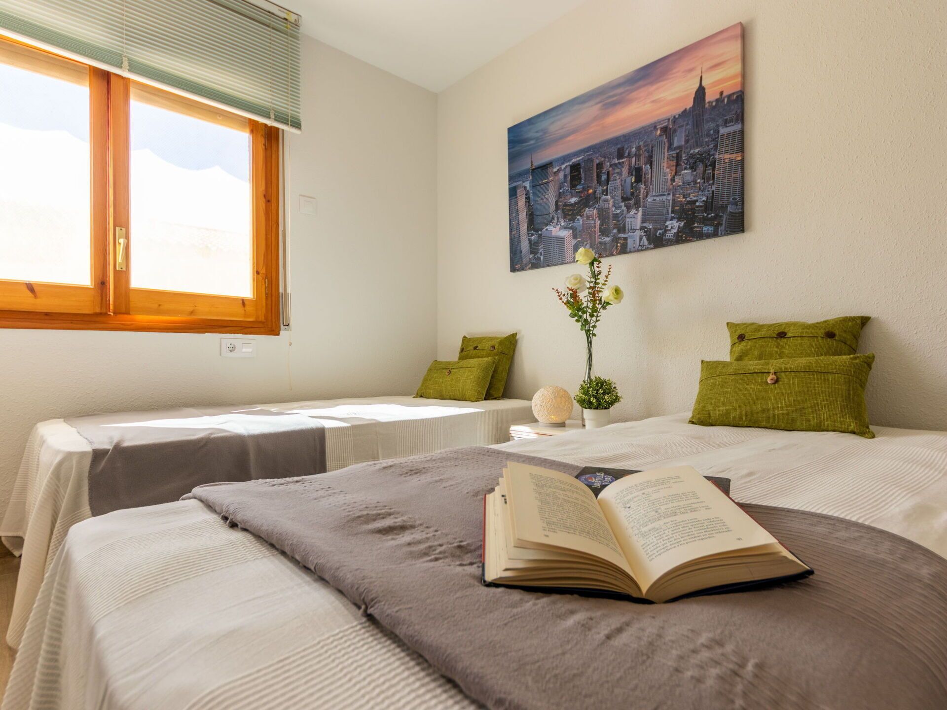 A hotel room with two beds, a window with blinds, a painting of a cityscape, and a small table with a vase and a book.