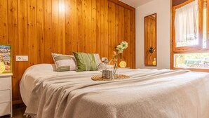 A bedroom with a wooden wall, a bed with a white bedspread, pillows, a bedside table with a lamp, and a window with curtains. - Wonderful private villa for 6 guests with WIFI, A/C, private pool, TV, patio and pets allowed (Deltebre)