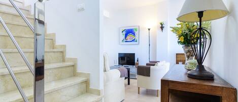 A modern living room with a staircase, a flat-screen TV, a sofa, and a lamp.