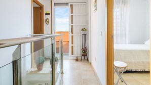 A hotel room with a bed, a small table, and a chair. There is a balcony with potted plants and a view of the sky.