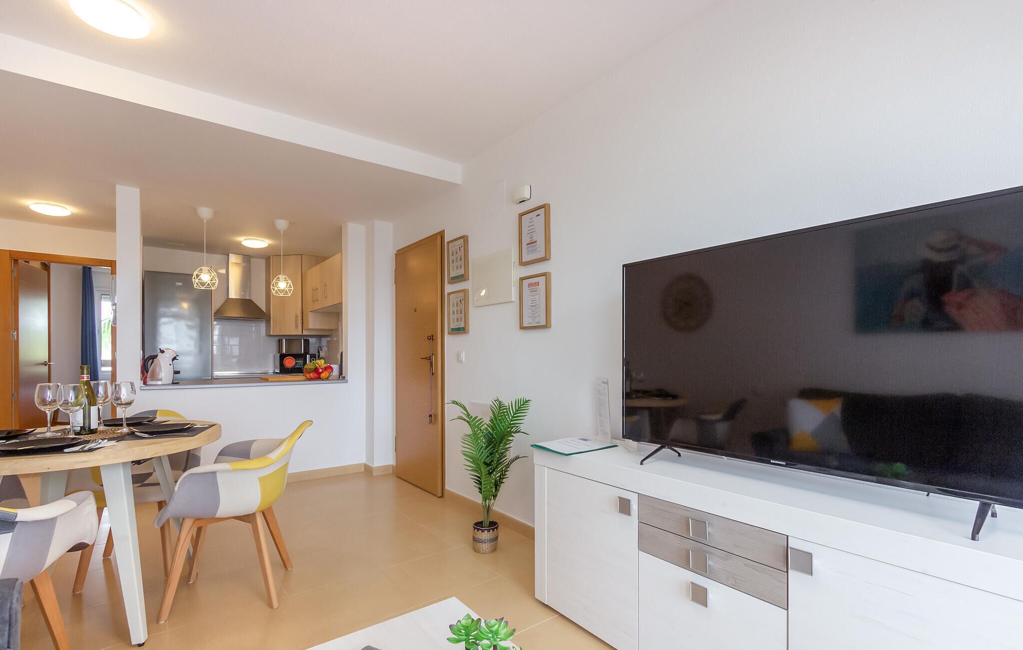 A modern living room with a flat-screen TV, a dining area with a table and chairs, a kitchen with cabinets and appliances, and a potted plant.