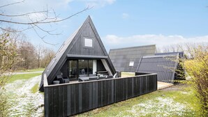 Ein modernes, dreieckiges Holzhaus mit einem großen Glasfenster und einer Terrasse mit Gartenmöbeln.