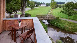 A wooden deck with a table and chairs, overlooking a river and a grassy field.